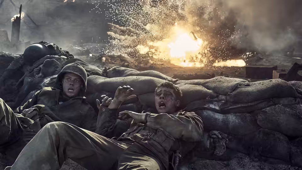 Two soldiers take cover behind sandbags as an explosion erupts nearby