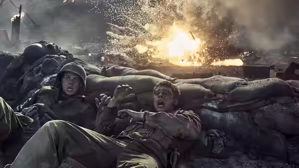 Two soldiers take cover behind sandbags as an explosion erupts nearby