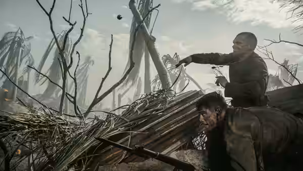 Two soldiers crouch beside a fallen tree amid a sparse woodland