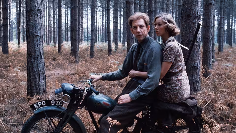 A man and woman ride a motorcycle through a sparse forest