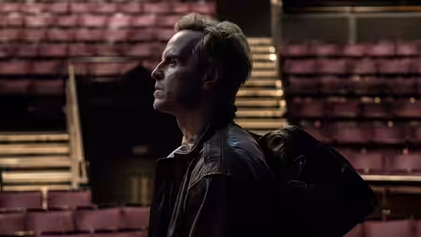 A man stands among rows of empty theatre seats, looking up toward the stage