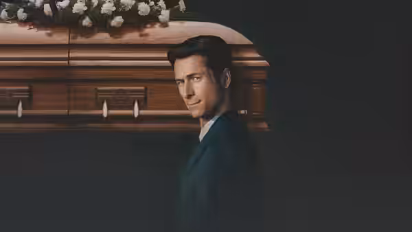 Becket (Glen Powell) standing beside a wooden coffin