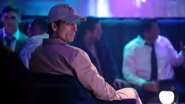 Becket (Glen Powell) seated in an audience wearing a cap