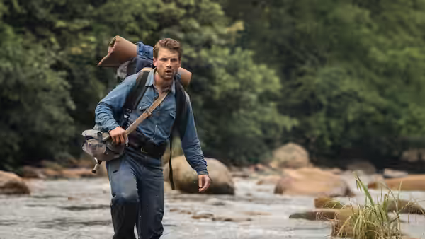 Yossi (Daniel Radcliffe) wades through a shallow jungle river carrying his pack