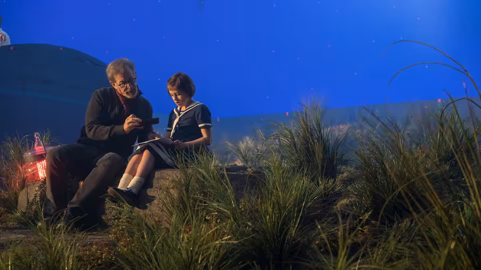 The BFG (Mark Rylance) sits beside Sophie (Ruby Barnhill) among tall grass beneath a blue sky