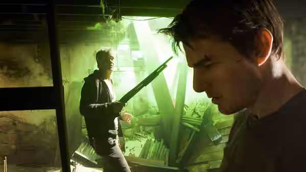 Robbie (Justin Chatwin) examines a weapon in a green-lit room as another man watches