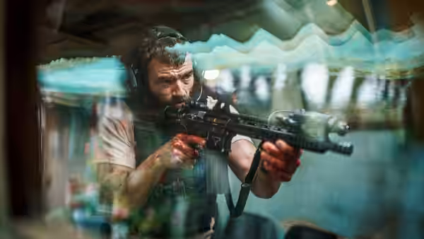 A bearded man aims a rifle behind reflective glass