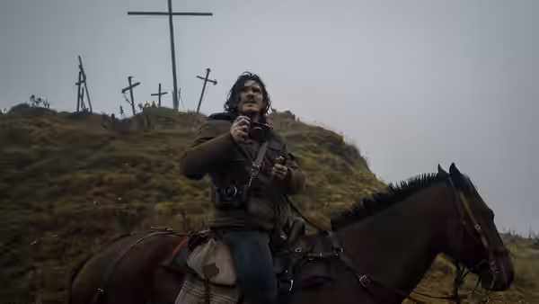 A long-haired man on horseback holds a camera near crosses on a hillside
