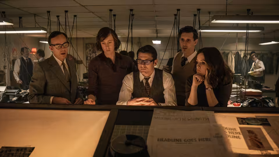 Five newspaper staff gather around a newsroom desk to review a broadsheet layout