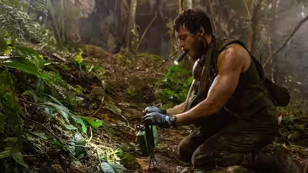 A soldier kneels in the forest, examining equipment beside a small green bag