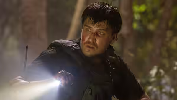 A man in dark tactical gear aims a flashlight through the forest