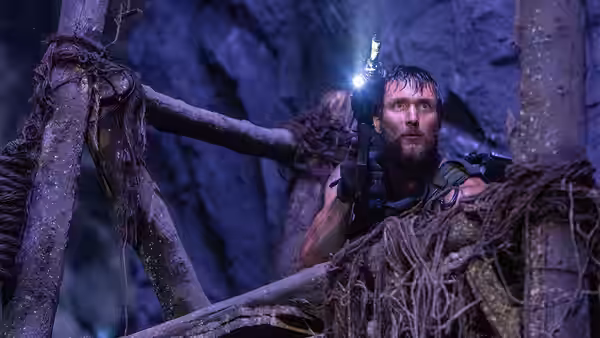 A bearded soldier peers through a tangled wooden structure, holding a rifle and light