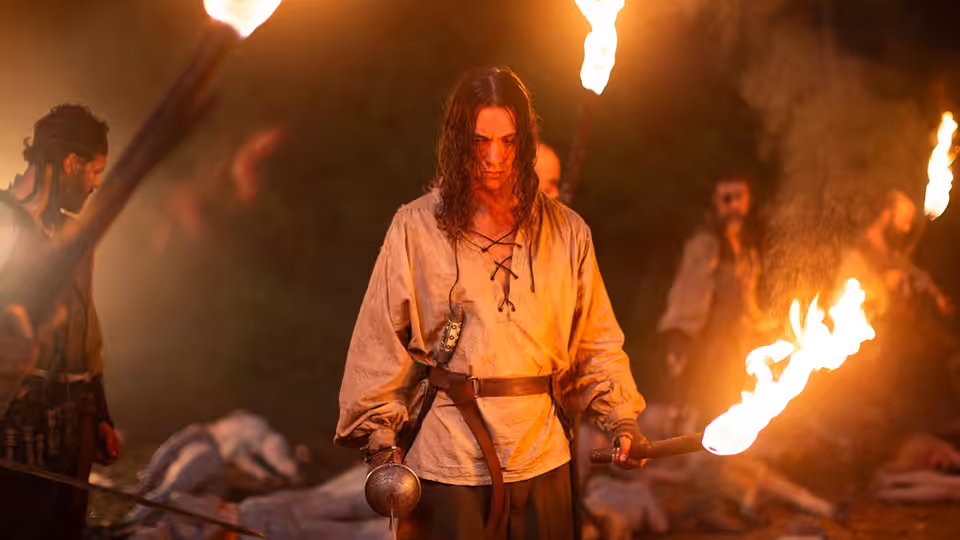A long-haired man holds a burning torch among bodies and other torchbearers