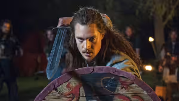 Uhtred of Bebbanburg (Alexander Dreymon) peers over a round shield with a sword raised behind him