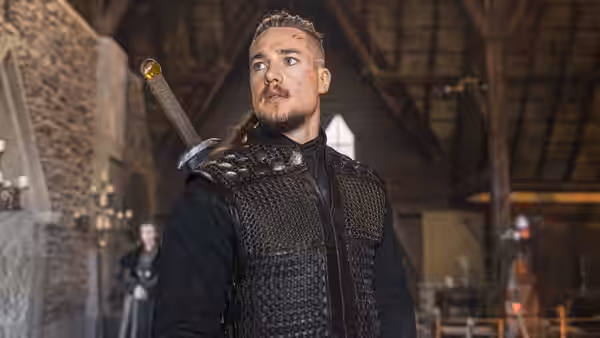 Uhtred of Bebbanburg (Alexander Dreymon) stands inside a timber hall with a sword on his back