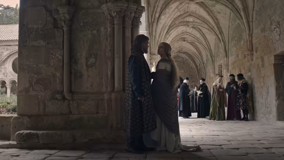 A man and woman stand beneath a stone archway as attendants gather in the corridor