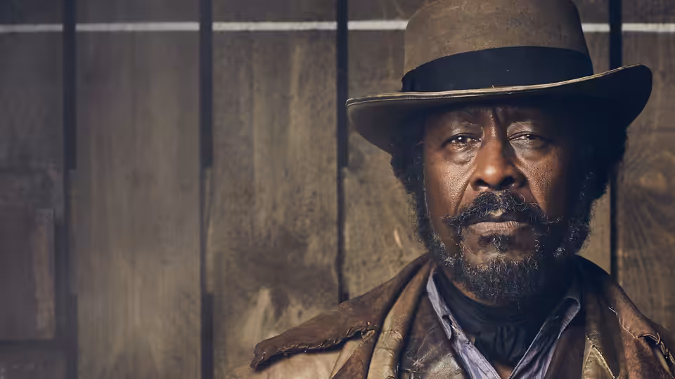 Ralph Coates (Clarke Peters) wearing a wide-brimmed hat indoors