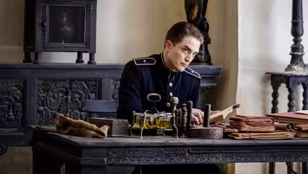 Officer Mandel (Robert Pattinson) examines objects and papers on a desk