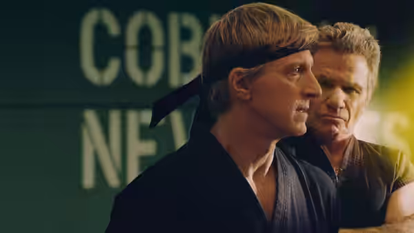 Johnny Lawrence (William Zabka) stands in profile before an older man and Cobra Kai lettering