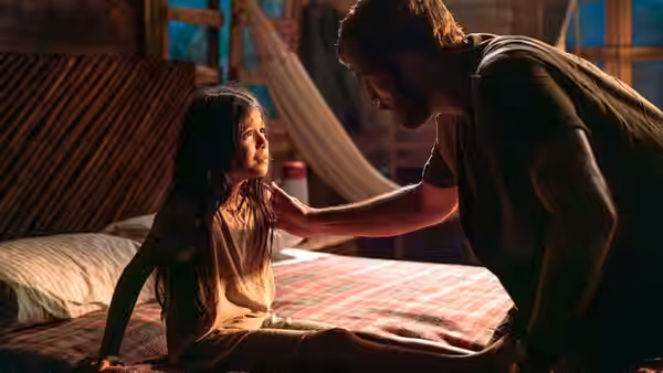 A man leans toward a seated girl on a bed and reaches for her shoulder