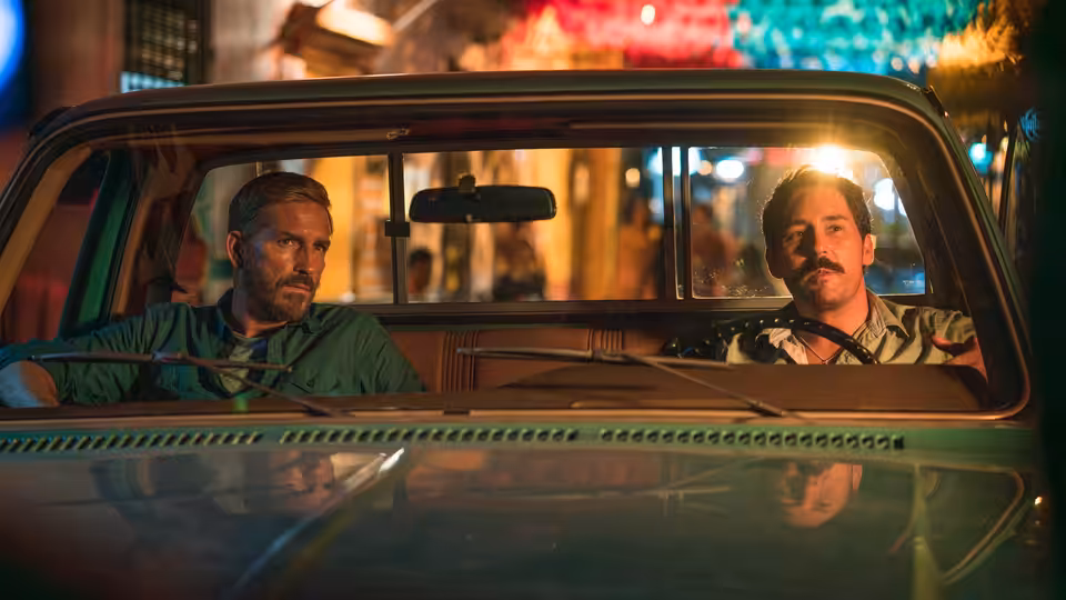 Two men sit in the front seats of a car parked among colorful city lights