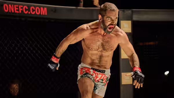 A bare-chested fighter in red gloves roars inside a fenced combat arena