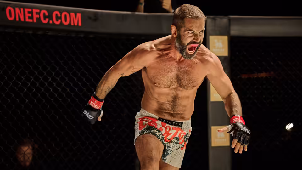 A bare-chested fighter in red gloves roars inside a fenced combat arena