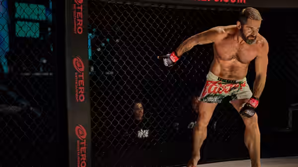 A bare-chested fighter in red gloves stands inside a fenced arena