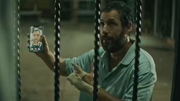 Stanley Sugerman (Adam Sandler) holds up a phone showing a smiling man, behind gym equipment