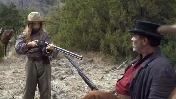 Brooke Briggs (Ryan Kiera Armstrong) aims a rifle toward a seated man beside a horse