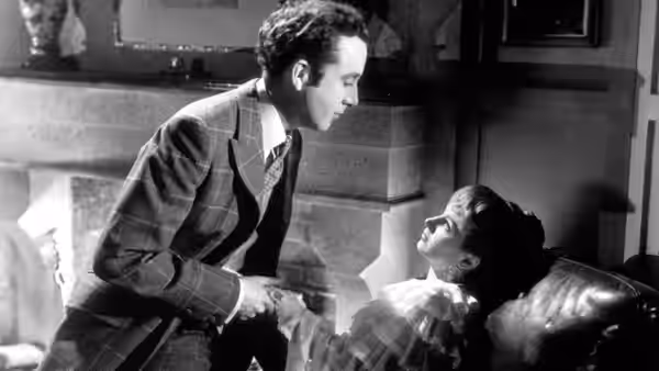 Dennis Price leans over a woman reclining on a couch by a fireplace