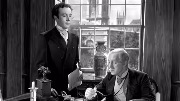 Dennis Price stands holding papers beside a seated older man at a desk