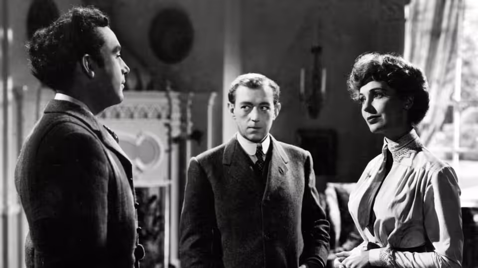 Dennis Price as Louis Mazzini stands between a man and a woman in a room