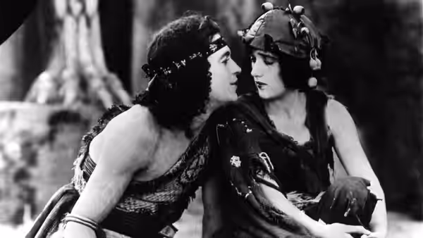 A woman in an ornate headdress leans close to another woman in a helmet