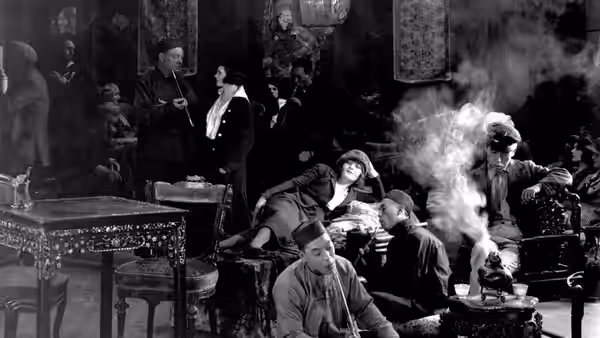 Men gather around tables in a smoky room as one man tends a pipe