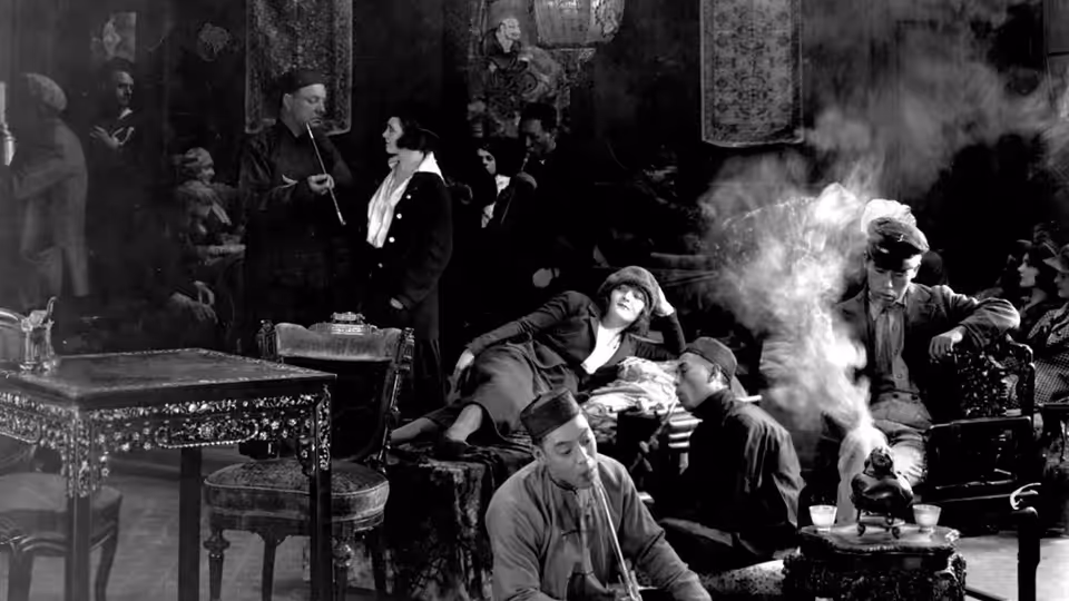 Men gather around tables in a smoky room as one man tends a pipe