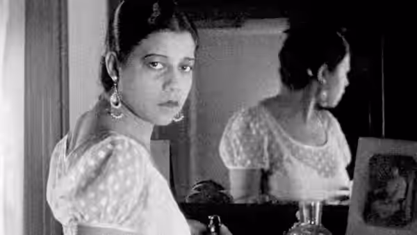 A woman in a light dress looks toward the viewer beside a mirror