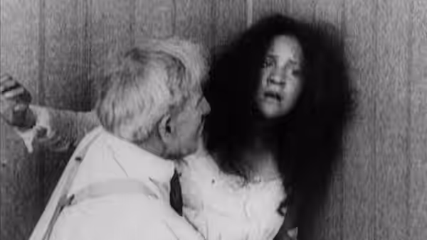 A seated older man reaches toward a woman standing beside him