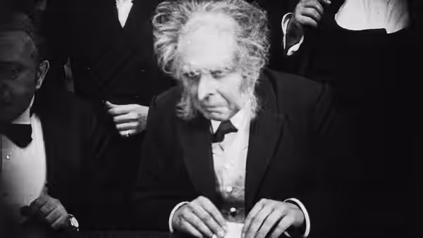 An older man bends over a document at a table as others stand behind him