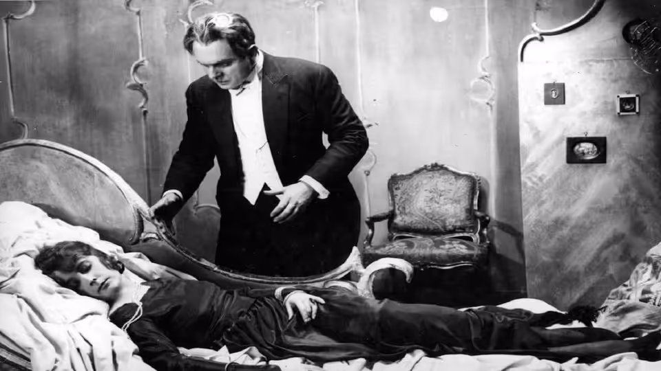 A suited man stands beside a bed where another man lies unconscious
