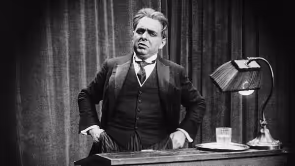 A suited man stands behind a desk beside a shaded lamp