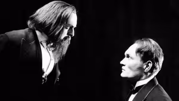 A bearded man leans toward a seated man against a dark background