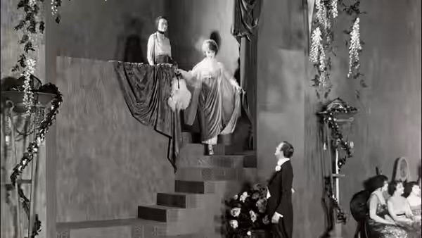A woman in a flowing gown descends a staircase as a man watches below