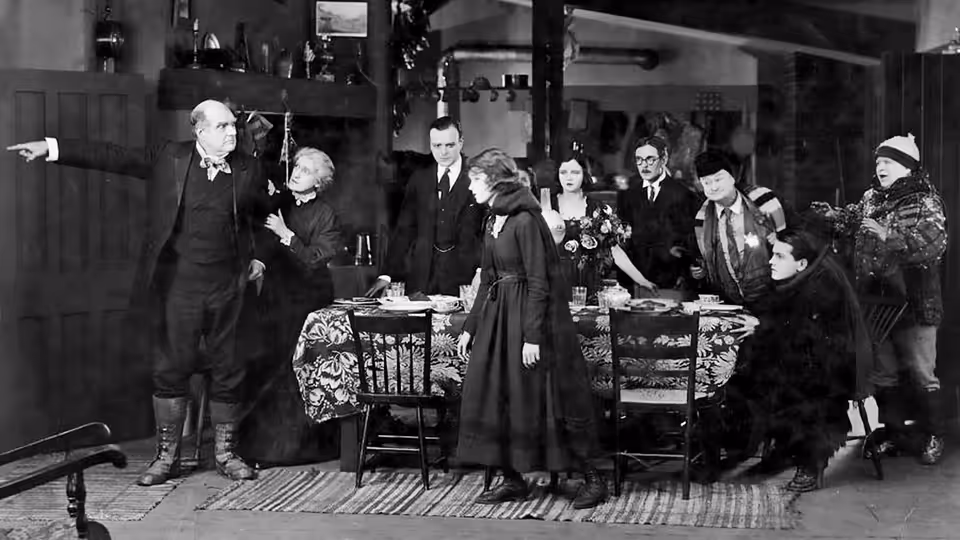 A woman stands beside a dining table as formally dressed guests gather around it