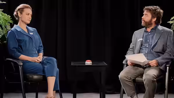 Carol (Lauren Lapkus) and Zach Galifianakis sit facing each other across an interview table