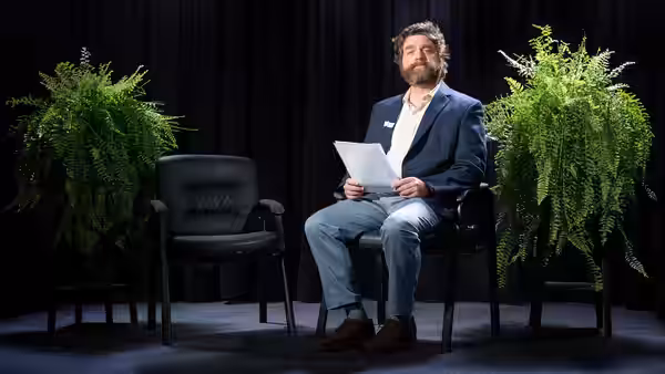 Zach Galifianakis sits holding papers between two large ferns