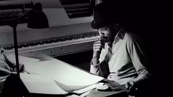 A man in a hat studies sheets of music at a desk beneath a lamp