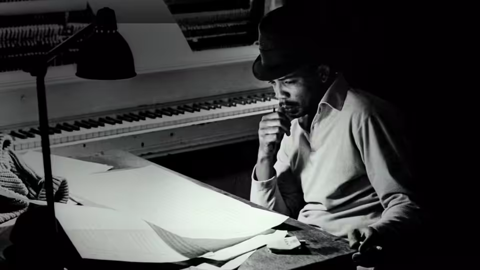 A man in a hat studies sheets of music at a desk beneath a lamp