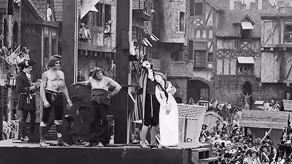 Henriette and Louise stand on a raised platform as a crowd gathers in the street