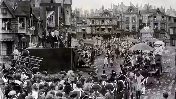 A crowd gathers in a town square around a raised platform and a large ship